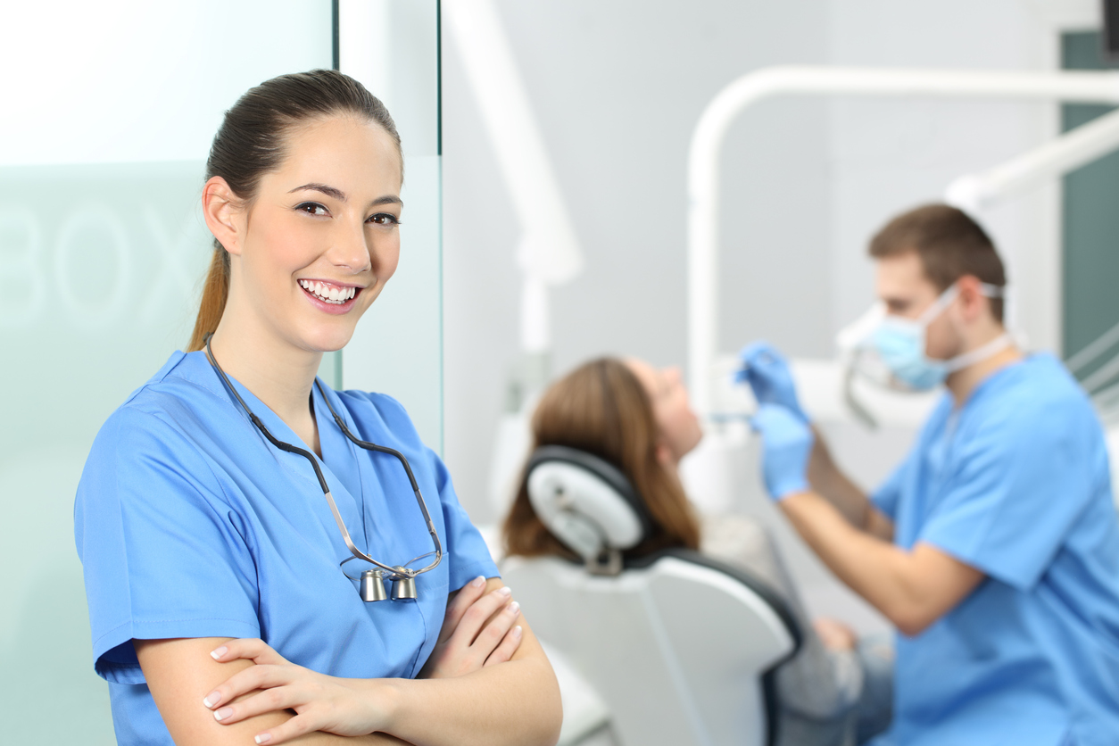 Dentist female with crossed arms wearing blue coat posing and looking at you at consultation with a doctor working with a patient in the background